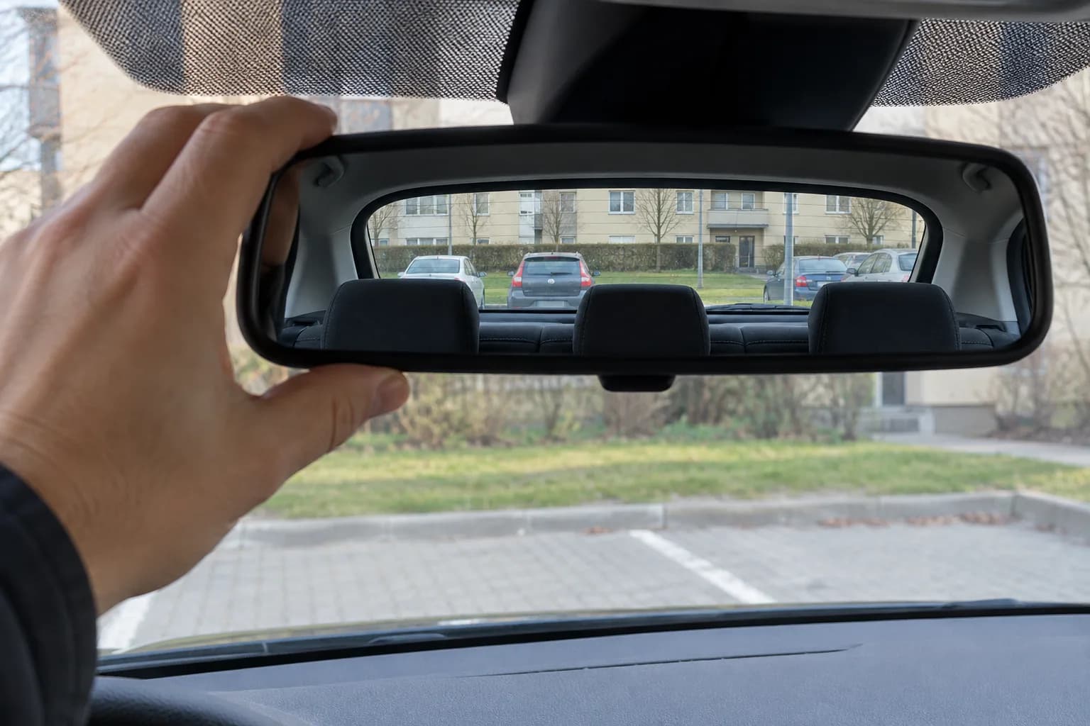 Dłoń kierowcy ustawia lusterko wsteczne w zaparkowanym aucie, w lusterku widać całą tylną szybę i parking pod blokiem.