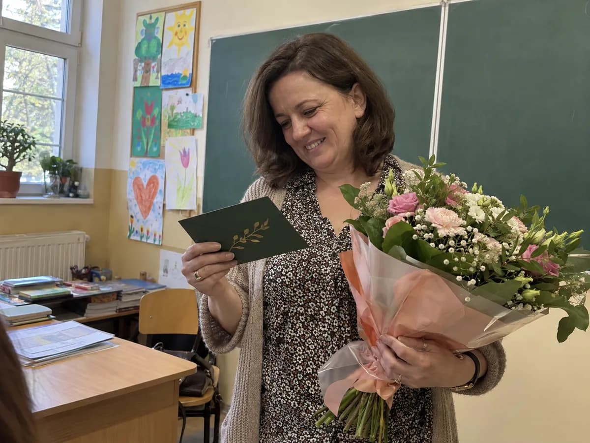 Nauczycielka przy biurku w szkolnej klasie, trzyma voucher na masaż w zielonej kopercie i uśmiecha się